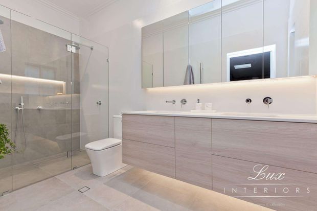 Bright, contemporary bathroom featuring a large frameless glass walk-in shower, wall-hung toilet, floating double vanity with pale wood-grain cabinetry, integrated basin, mirrored wall cabinets, concealed LED lighting, and large neutral stone-look floor and wall tiles.