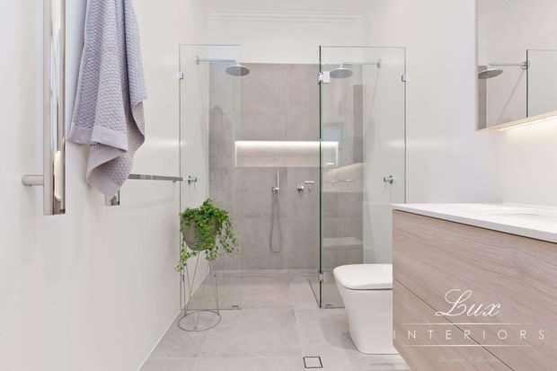 Bright contemporary bathroom with a walk-in glass shower, wall-mounted rain shower fixtures, freestanding toilet, pale tiled walls and floor, and a light timber-look floating vanity with a white benchtop. A towel rail, folded towel, small plant, and illuminated mirror add subtle detail.