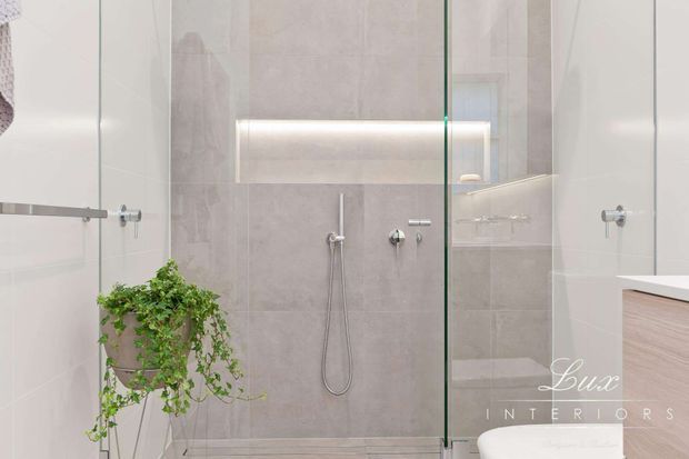 Bright contemporary bathroom with a walk-in glass shower enclosure, pale large-format wall and floor tiles, concealed shower fittings, recessed illuminated niche, floating timber-toned vanity, and a trailing green plant.