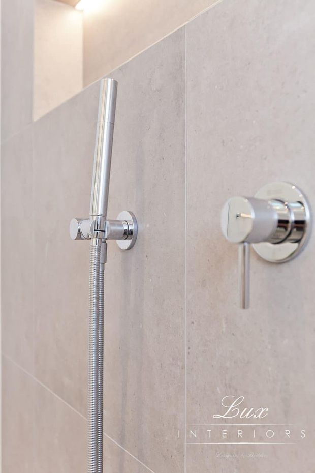 Close-up of a minimalist wall-mounted handheld shower and chrome control set against large-format pale beige stone-look bathroom tiles. The polished chrome fixtures and clean grout lines create a refined, contemporary bathroom detail.