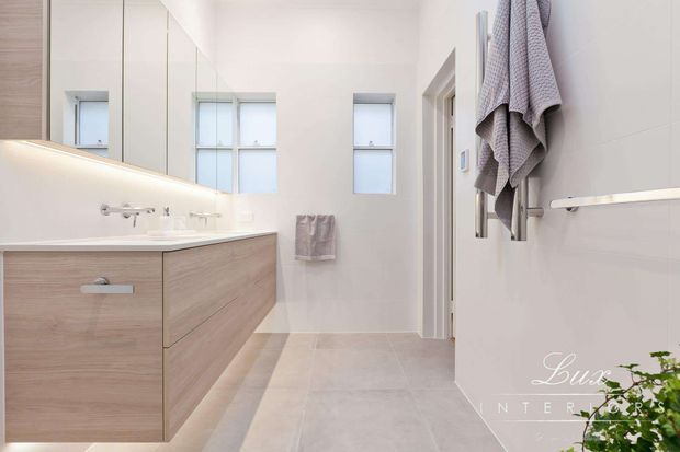 Bright contemporary bathroom featuring a long floating timber-look vanity with integrated basin, large mirrored wall cabinets, pale tiled flooring and walls, multiple windows, and wall-mounted towel rails. The room has a clean, minimalist fit-out with warm wood cabinetry and soft grey textiles.