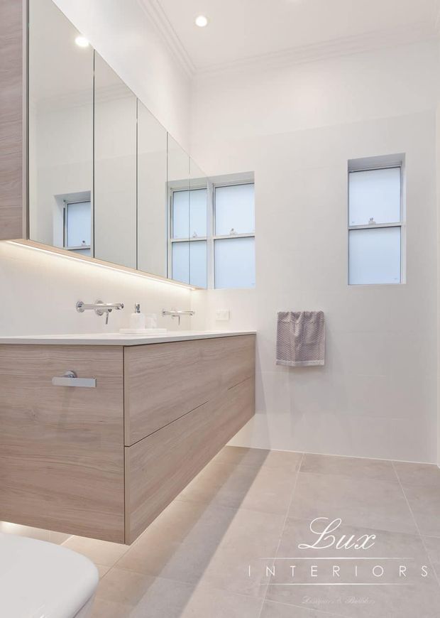 Bright contemporary bathroom featuring a floating light timber vanity with integrated drawers, pale stone-look tiled flooring, white walls, a large mirrored cabinet, dual wall-mounted basins and chrome tapware. Frosted windows provide natural light, while concealed vanity lighting and recessed ceiling lights create a clean, refined atmosphere.