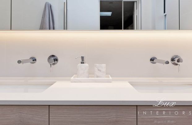 Close-up of a refined contemporary bathroom vanity with a wide pale stone or solid-surface benchtop, wall-mounted chrome tapware, integrated basin area, soap and tissue accessories, warm concealed under-mirror lighting, and grey wood-grain cabinetry. A large mirror reflects a towel and part of the room.
