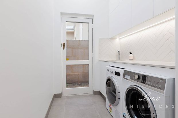 Bright, compact laundry room with white built-in cabinetry, a white herringbone-tile splashback, grey floor tiles, and side-by-side front-loading washing machine and dryer. A glazed exterior door provides natural light, and under-cabinet lighting illuminates the benchtop.