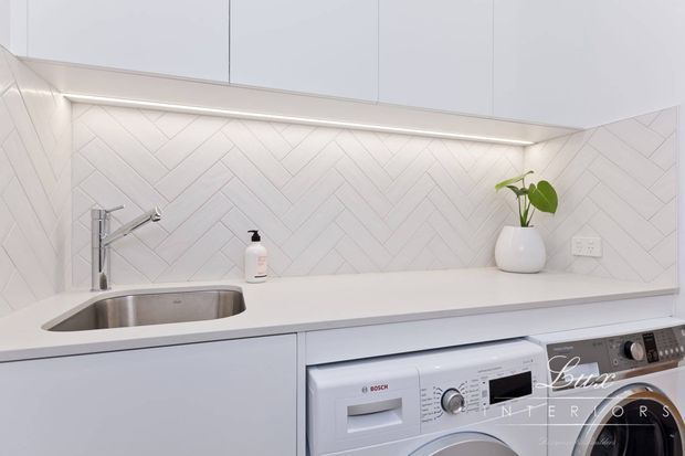 Bright, compact contemporary laundry and utility area with white handleless overhead cabinetry, a white engineered stone benchtop, stainless-steel sink and tap, and a white herringbone tiled splashback. An integrated front-loading washing machine and adjacent appliance sit beneath the benchtop, with subtle under-cabinet lighting and a small potted plant adding warmth.