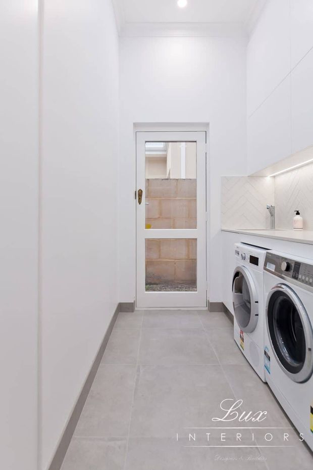 A narrow, bright contemporary laundry room with white cabinetry and walls, a pale grey tiled floor, a white herringbone tile splashback, under-cabinet lighting, and a front-loading washing machine and dryer beneath a long benchtop. A glazed door at the end opens toward an exterior or utility area.