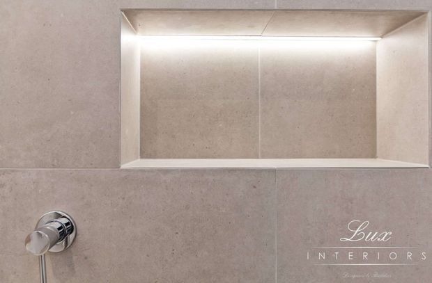 Close-up of a minimalist tiled bathroom wall featuring a recessed rectangular niche with integrated concealed lighting and a chrome wall-mounted fixture. The niche and surrounding wall are finished in a warm light beige stone-look surface.