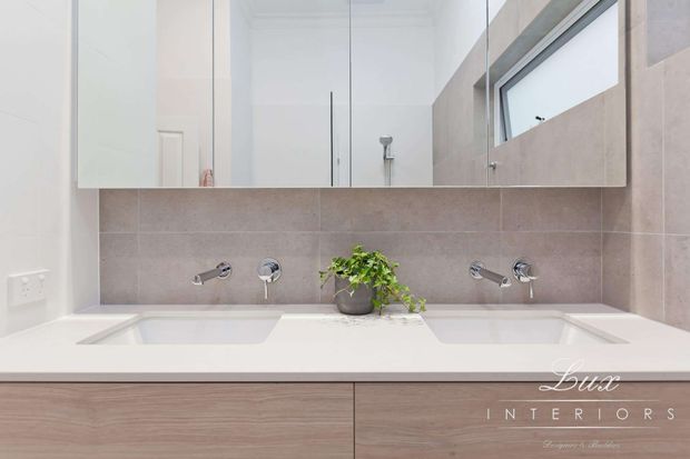 Bright contemporary bathroom vanity with a wide double basin in a pale engineered stone benchtop, warm grey wall tiles, light timber-look cabinetry, wall-mounted chrome tapware, and a large mirrored cabinet. A high horizontal window provides natural light, while a small potted plant adds greenery.