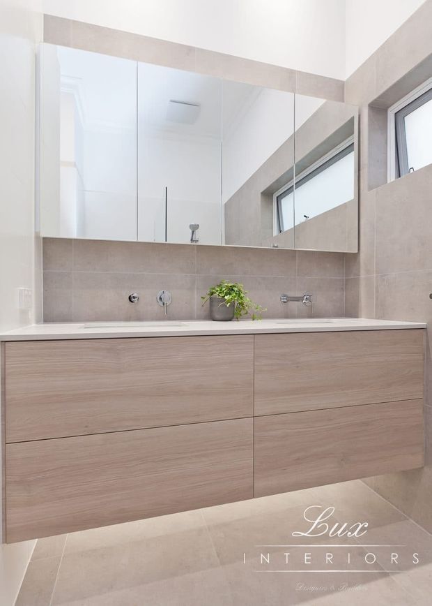 Bright contemporary bathroom vanity with a wall-to-wall floating timber-look cabinet, white benchtop, large mirrored shaving cabinet, neutral beige tiled walls and floor, chrome wall-mounted tapware, and a small potted plant. High-set windows provide natural light.