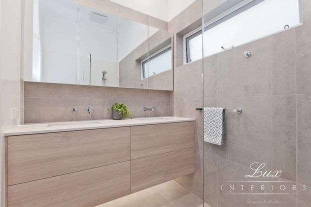 Bright contemporary bathroom featuring a long floating timber-look vanity with integrated countertop, wall-to-wall mirror, large-format warm grey-beige wall and floor tiles, and a frameless glass shower enclosure. High horizontal windows provide natural light, while minimal chrome fixtures and a small plant add restrained detail.