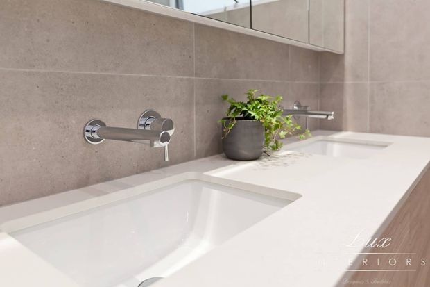 Contemporary bathroom vanity with a long white built-in countertop, two integrated rectangular basins, wall-mounted chrome faucets, large-format taupe-grey wall tiles, and mirrored cabinetry above. A small potted plant adds a natural accent.