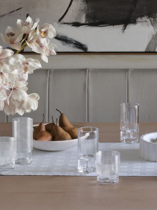 A styled dining table vignette with a pale wood tabletop, upholstered vertical-panel banquette, ribbed clear glassware, a white serving bowl filled with pears, white orchids, and a large abstract black-and-white artwork behind the seating. The scene has a refined, neutral contemporary aesthetic with soft grey, white, beige, and brown tones.