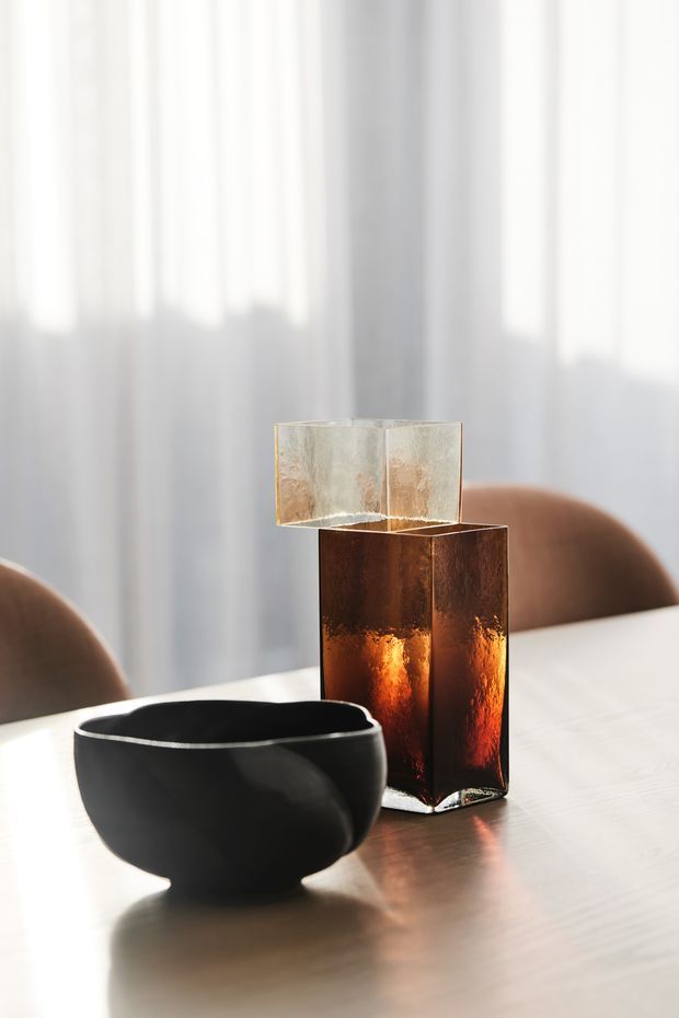 A softly lit dining setting featuring a matte black sculptural bowl and a tall amber-brown translucent glass candleholder or lamp on a pale wooden table. Warm upholstered dining chairs and sheer white curtains create a refined, calm atmosphere with gentle natural light.