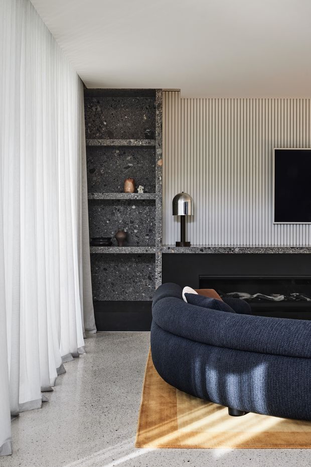 A refined contemporary living room with a curved deep-blue sectional sofa facing a dark linear fireplace and wall-mounted television. The room features a dramatic dark stone open shelving surround, a matching stone fireplace ledge, vertically ribbed light wall panelling, polished metal table lamp, full-height sheer curtains, and a pale terrazzo floor. Strong natural light creates a calm, luxurious atmosphere.