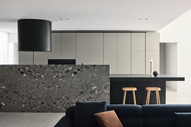 A refined open-plan kitchen and living area with a large dark terrazzo-style stone island, pale grey full-height cabinetry, black benchtops, a sculptural black rangehood, and timber bar stools. A dark sofa with a warm brown cushion occupies the foreground. The space has clean contemporary lines, recessed lighting, and a restrained neutral palette.