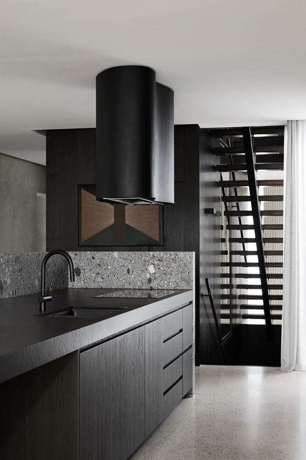 A moody, contemporary kitchen with dark textured cabinetry, a black stone benchtop, and a large terrazzo or concrete-style splashback. A sculptural black cylindrical rangehood hangs above the island, with a black sink and tap in the foreground. Behind the kitchen is a dark timber-clad wall with a recessed decorative feature, while an open-riser staircase with dark framing leads upward beside sheer curtains. The space has a refined monochromatic palette and polished concrete or terrazzo flooring.