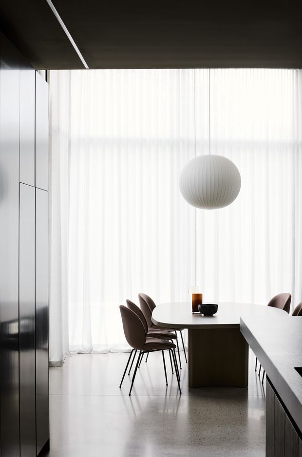 A refined, minimalist dining area integrated with a kitchen. A round white dining table is surrounded by dark upholstered chairs, with a sculptural ribbed pendant light suspended in front of sheer floor-to-ceiling curtains. Dark built-in cabinetry lines the left wall and a large dark kitchen island or counter occupies the foreground. The space has a polished pale floor, dark ceiling, and a restrained monochromatic palette.