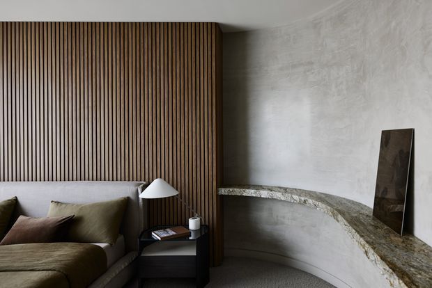 A refined living room with a low sectional sofa, layered neutral cushions, a black side table and sculptural table lamp. Vertical timber battens form a feature wall behind the sofa, while a curved stone ledge wraps around a textured grey plaster wall and meets a dark rectangular artwork. The palette is warm, muted and sophisticated.