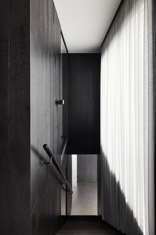 A narrow, dramatic interior stairway descends between dark timber-clad walls and a full-height sheer-curtained window. A slim black handrail runs along the wall, with a bright lower-level landing visible beyond. The composition emphasizes strong vertical lines, deep shadows, and filtered natural light.
