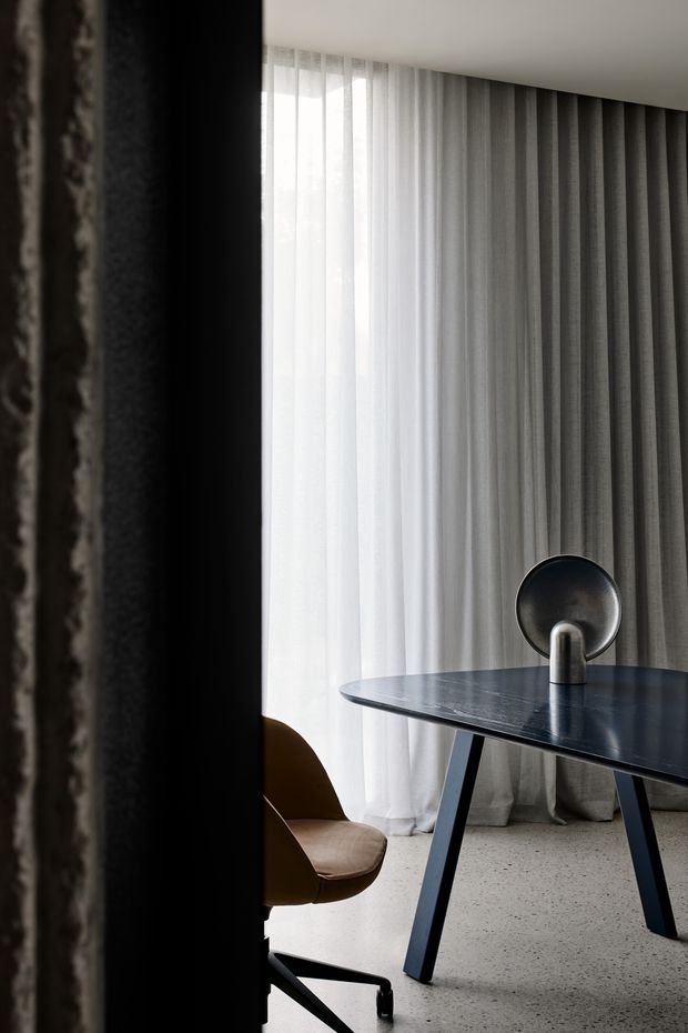 A moody, minimalist interior vignette featuring a dark rectangular table with angled legs, a sculptural circular-backed lamp or accessory, and a brown upholstered office chair. Floor-to-ceiling sheer white and grey curtains provide soft diffused light, while a dark textured wall or structural column frames the left side. The floor appears to be light terrazzo or speckled concrete.