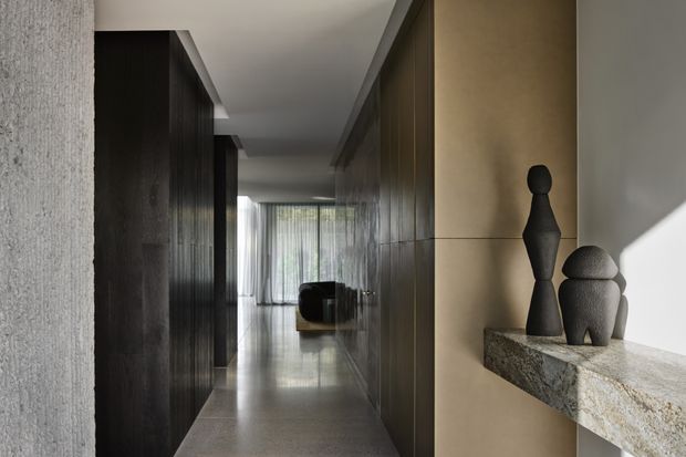 A refined contemporary hallway with dark timber wall panelling and cabinetry on the left, warm beige feature walls, polished stone flooring, and a projecting natural-stone display shelf holding sculptural objects. The corridor leads toward a glazed living area with sheer curtains and soft natural light.
