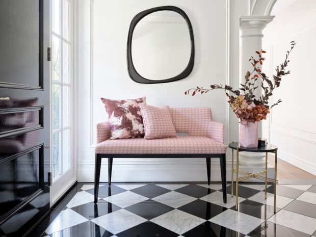 A refined entryway or transitional sitting area with a pale pink upholstered bench, patterned cushions, black-and-white checkerboard flooring, white wall panelling and moulded architectural details. A glossy black built-in cabinet or door occupies the left side, while a slim brass-and-black side table holds a tall floral arrangement. A rounded black-framed mirror adds a bold graphic accent.