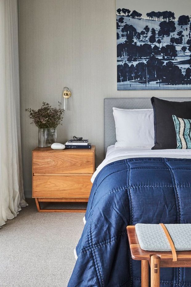 A refined bedroom corner featuring a grey upholstered bedhead, white bedding, and a deep navy quilt. A warm timber two-drawer bedside table sits beside the bed with a glass vase, books, and a small brass-toned lamp. Full-height sheer curtains, a textured neutral wall, and a monochrome landscape artwork complete the calm, tailored scheme. A timber bench with a light cushion is visible at the foot of the bed.