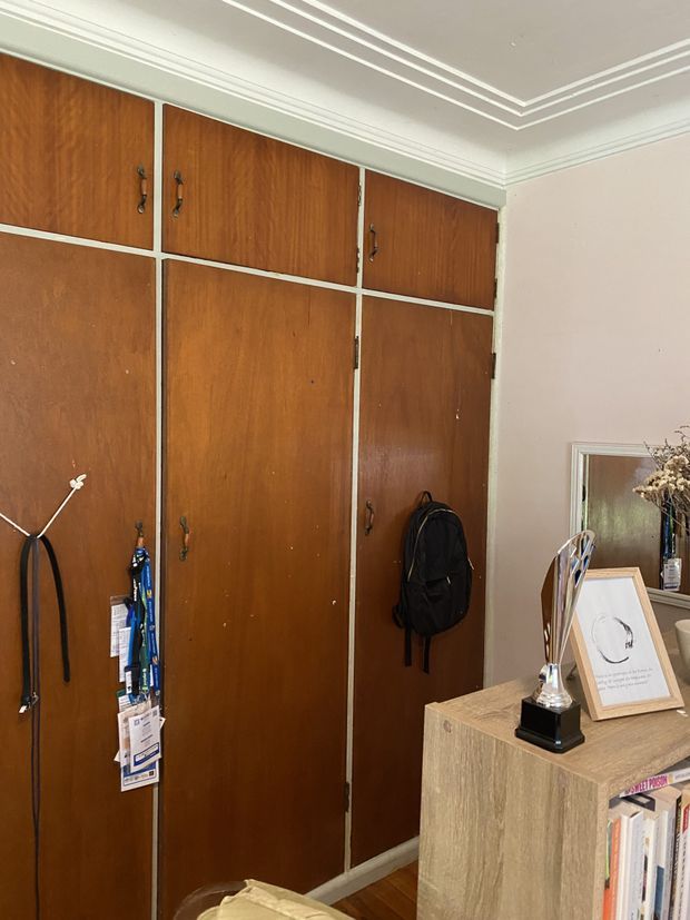 Interior view of a traditional timber-lined storage wall with tall paneled cupboard doors and dark metal handles. A wood-toned console or bookcase with books, framed artwork, and a trophy occupies the foreground, while a framed mirror and decorative objects appear at right. The room has a light painted ceiling with layered decorative cornice moulding and timber flooring.