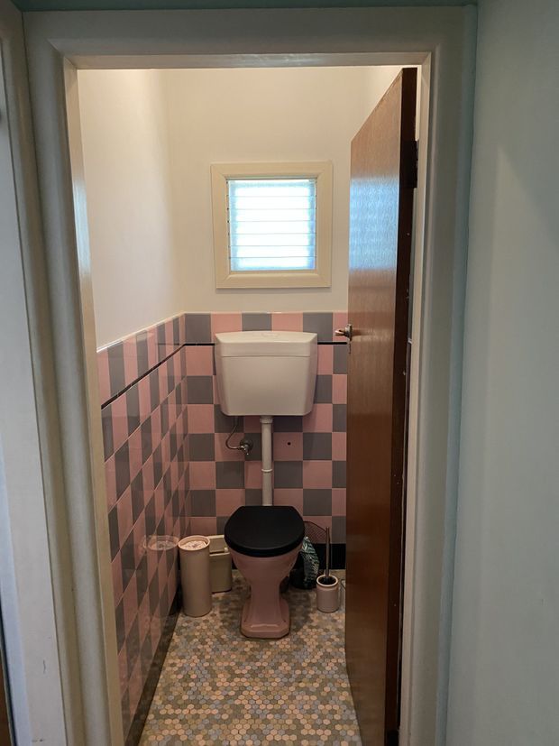 A compact separate toilet room viewed through its open doorway. The room features a wall-mounted cistern and toilet, pink and dark grey checkerboard wall tiles, a small patterned mosaic floor, a high horizontal louvre window, painted walls, and a timber door.