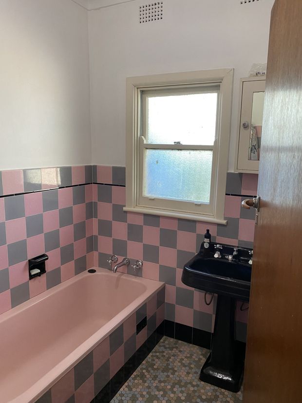 Compact vintage bathroom with a pink bathtub, black pedestal basin, and retro pink, grey, and black checkerboard wall tiles. The room has a frosted sash window with painted timber trim, a small patterned mosaic floor, and a wooden door.