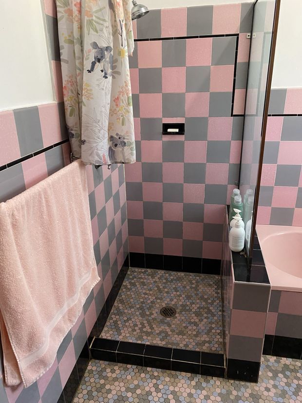 A compact, retro-style bathroom with a walk-in shower enclosed by clear glass. The shower and surrounding walls feature a bold pink and grey checkerboard tile pattern with black grout and black tiled trim. The shower floor uses small multicoloured hexagonal mosaic tiles, coordinated with the bathroom floor. A floral shower curtain, pink towel, toiletries, and part of a pink bathtub are visible.