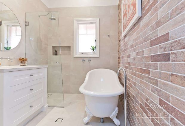 Bright traditional bathroom featuring a freestanding clawfoot bathtub, frameless glass shower, white vanity, round wall mirror, pale stone-look wall and floor tiles, and a red-brown exposed brick feature wall.