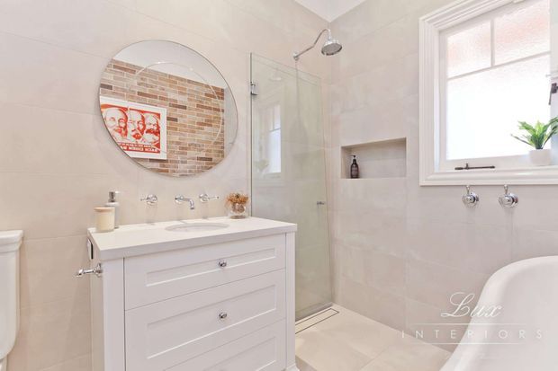 Bright, refined bathroom with a white vanity and integrated basin, wall-mounted tapware, frameless glass shower enclosure, recessed wall niche, pale tiled walls and floor, and a traditional sash-style window. A round mirror reflects a warm brick feature wall and artwork, while a freestanding bathtub is partially visible at right.