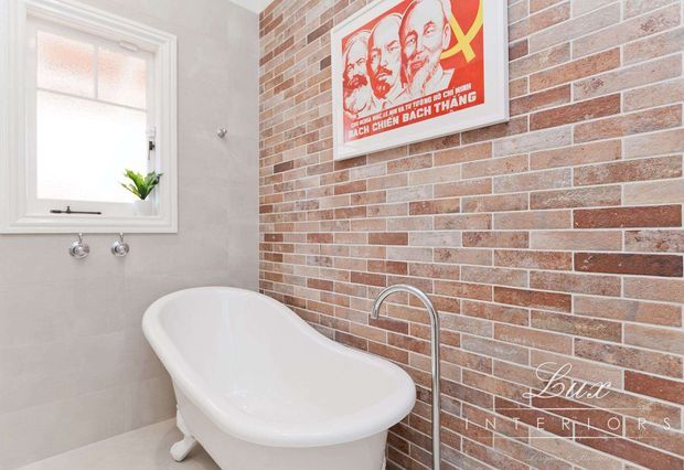Bright bathroom featuring a freestanding white clawfoot-style bathtub, white-framed frosted window, chrome wall taps and floor-mounted bath filler. One wall is clad in exposed red and cream brick tiles, contrasted with pale grey tiled walls and flooring. A framed graphic artwork hangs above the bath.