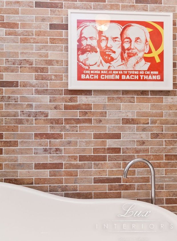 Close-up interior detail of a freestanding white bathtub with a tall curved chrome faucet, set against a warm reddish-brown exposed brick wall. A framed red political poster provides a bold decorative accent above the tub.