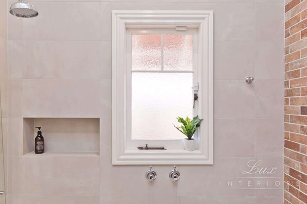 Bright traditional-style bathroom with pale tiled walls, a framed frosted-glass window, wall-mounted bath or shower controls, a recessed shower niche, and a small potted plant on the sill. A round showerhead is visible at the upper left, while exposed brickwork forms a contrasting wall edge on the right.