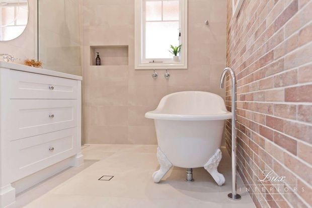 A bright, elegant traditional bathroom featuring a freestanding white clawfoot bathtub, white vanity cabinetry, pale beige tiled walls and floor, a recessed wall niche, chrome fixtures, and a contrasting exposed brick feature wall.