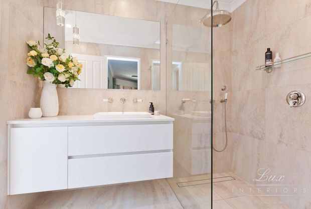 Bright, refined contemporary bathroom featuring a floating white double-drawer vanity, large frameless mirror, pale stone-look wall and floor tiles, wall-mounted basin fittings, and a frameless glass walk-in shower with rainfall showerhead and recessed shelf. Decorative pendant lights and a floral arrangement add a soft, elegant touch.