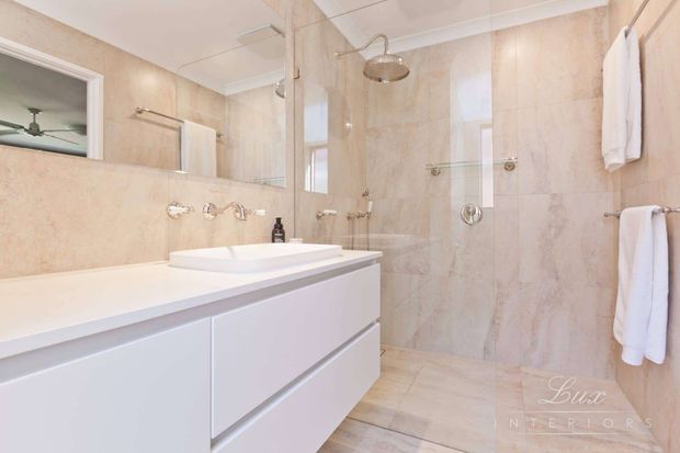 Bright, refined bathroom featuring a white floating vanity with integrated basin, large frameless glass shower enclosure, wall-mounted shower fittings, and full-height beige stone-look wall and floor tiling. White towels and polished chrome fixtures provide subtle contrast.