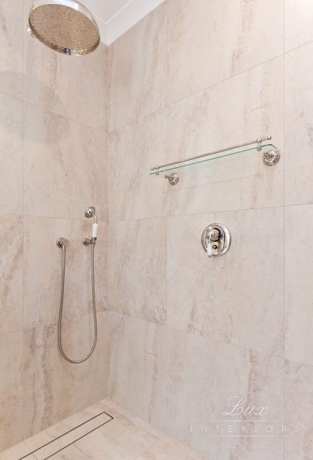 A minimalist walk-in shower with full-height pale beige stone-look wall and floor tiles, a large round rainfall showerhead, handheld shower, chrome controls, and a frameless glass shelf. The space is bright, neutral, and refined with a linear floor drain.