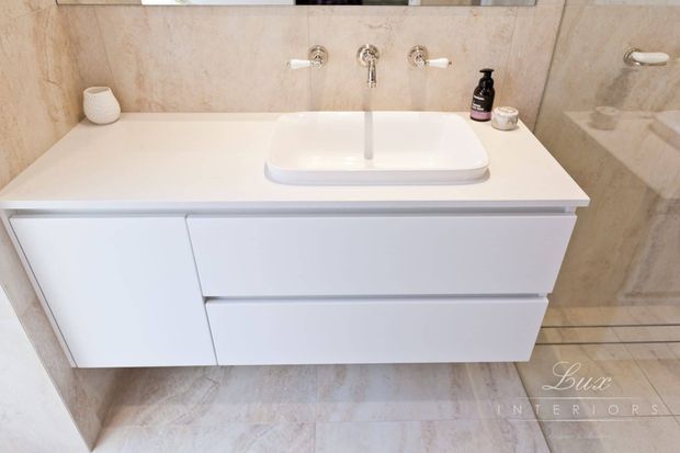 A close-up view of a refined bathroom vanity featuring a wall-mounted white handleless cabinet, white engineered-stone benchtop, and a rectangular vessel basin. Polished chrome wall-mounted tapware contrasts with beige natural-stone-look wall and floor tiles. A large mirror and subtle bathroom accessories complete the clean, contemporary scheme.