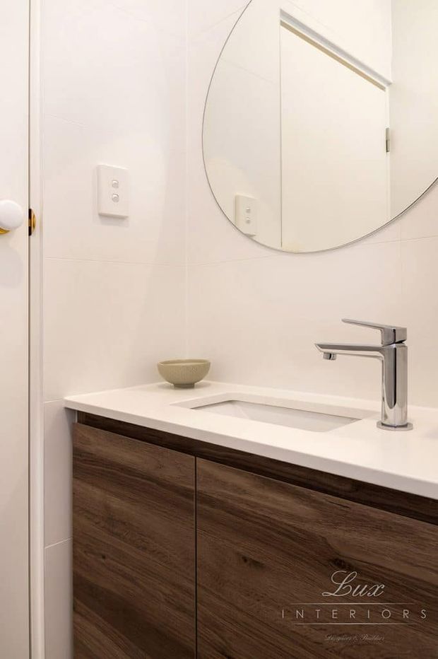 A compact, clean-lined bathroom vanity with a white engineered stone benchtop and integrated rectangular basin, paired with a chrome single-lever tap. The vanity has dark timber-look fixed cabinetry, while pale tiled walls, a large round mirror and a simple wall switch create a bright, minimalist interior.