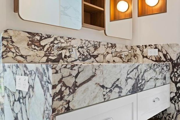 Close-up of a refined bathroom vanity with dramatic white and dark-veined natural stone wrapping the benchtop, backsplash, and side panels. The vanity has white drawers with chrome pulls, chrome wall-mounted fixtures, rounded white cabinetry, warm timber open shelving, and recessed circular lights.