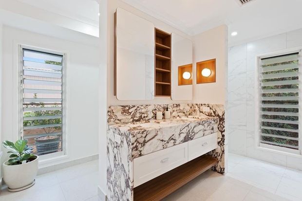 Bright contemporary bathroom featuring a sculptural double vanity wrapped in dramatic veined stone, white integrated cabinetry with open timber shelving, warm wood storage below, and square illuminated wall niches. White tiled walls and floors, plantation-style louvre windows, and a potted plant create a clean, airy feel.