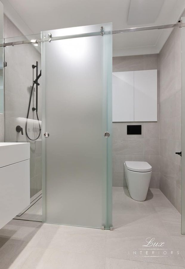 Bright contemporary bathroom with large pale grey tiled walls and floor, a frameless frosted-glass sliding shower screen with exposed chrome hardware, a wall-mounted white vanity, black shower fittings, concealed cistern cabinetry and a modern white toilet. The space has clean lines and a restrained neutral palette.