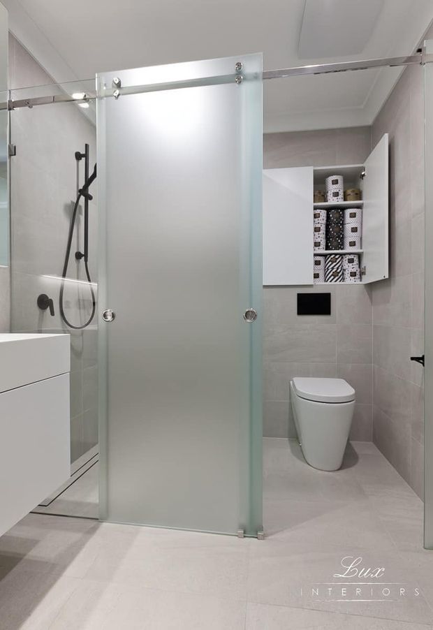 Bright contemporary bathroom featuring pale grey tiled walls and floor, a frameless frosted-glass sliding shower screen with exposed metal hardware, wall-mounted shower fittings, a white floating vanity, and a white toilet. Open white cabinetry provides storage above the toilet.