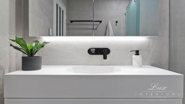 Contemporary bathroom vanity with a wall-mounted white basin and black tapware, set against large-format light grey wall tiles. A wide mirror with integrated underlighting reflects a towel rail and shower area, while a small plant and soap dispenser add restrained styling. The space has a clean, minimalist, neutral appearance.