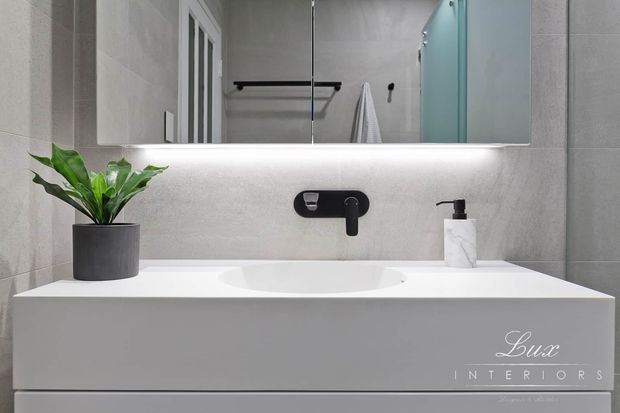 A contemporary bathroom vanity with a white integrated basin and wall-mounted matte black tapware. Large grey stone-look wall tiles, a frameless mirrored cabinet, under-cabinet lighting, black hardware, and a small potted plant create a clean, refined fit-out.