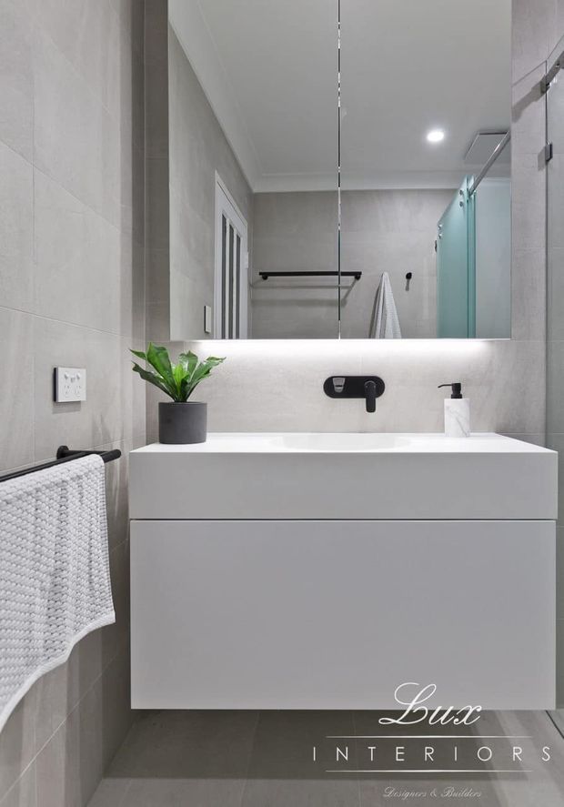 Contemporary bathroom vanity area with a wall-mounted white double-drawer cabinet, integrated white basin, matte black wall-mounted tapware, large mirror with concealed lighting, pale grey tiled walls, and a frameless glass shower enclosure. A small potted plant and textured white hand towel add restrained accents.