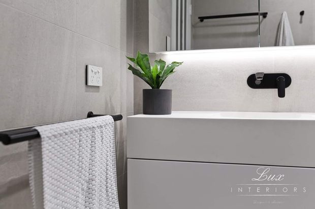 Close-up of a contemporary bathroom vanity with a white floating cabinet, integrated basin, and matte black wall-mounted tapware. Large pale grey wall tiles, a black towel rail with a textured white towel, mirrored cabinetry, and a small potted plant create a clean, restrained look.